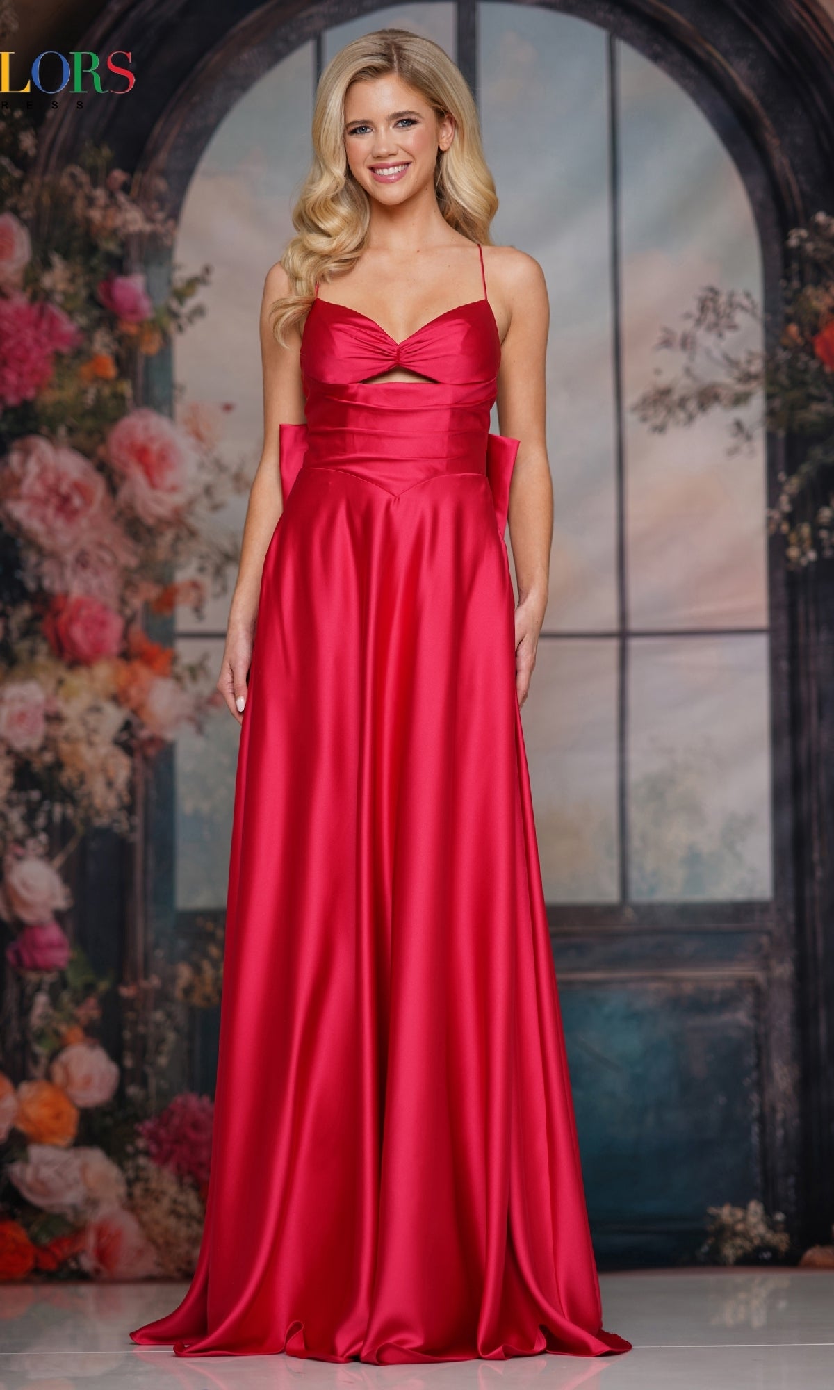 A woman wears the Colors Dress 3851 Back-Bow Strappy Prom Gown in Lipstick, a sleeveless floor-length satin evening dress, posing before a floral backdrop and large arched window.
